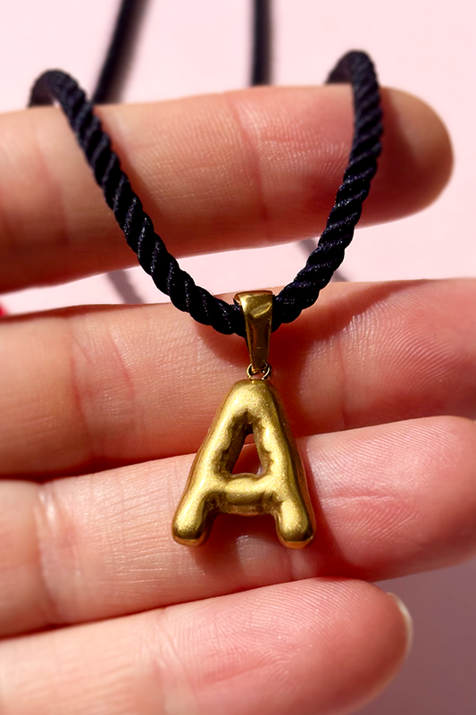 Gold 'A' pendant on a black cord held between fingers against a pink background