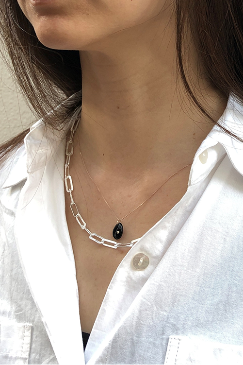 A close-up of a woman wearing black cat-eye sunglasses, a white button-up shirt, and layered silver necklaces against a textured beige wall.