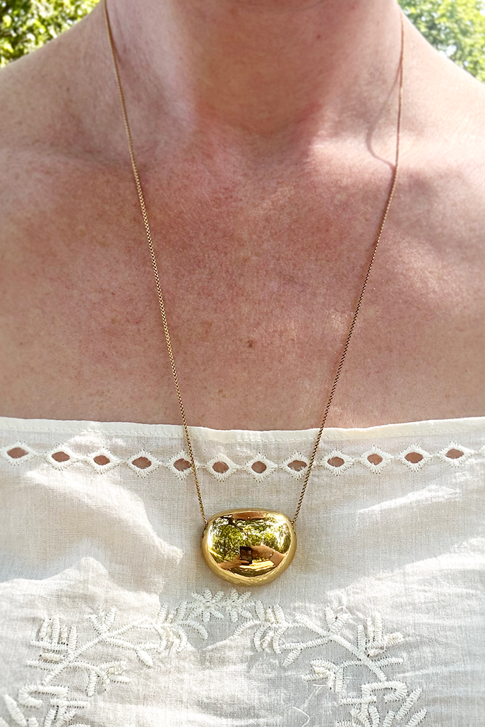 A close-up of a person wearing a delicate gold chain necklace with a polished, rounded gold pendant, styled over a white embroidered top.